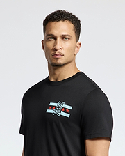 A man with short curly hair is wearing the Psycho Bunny MENS CHICAGO TEE - B6U551W1PC, crafted from soft Pima cotton, showcasing a rabbit skull and crossbones graphic over a stylized flag with red stars and blue stripes. The background is plain white.