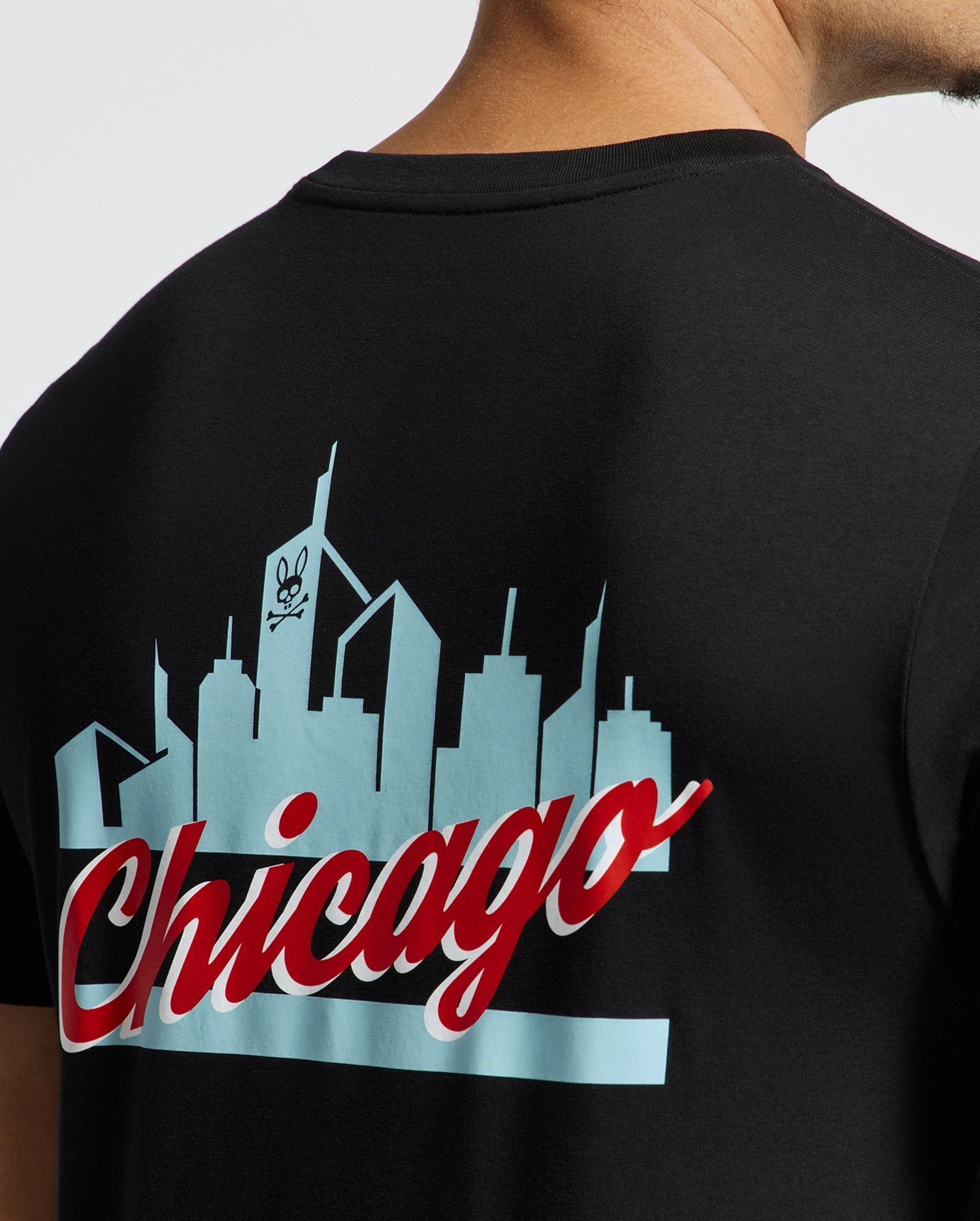 A person is wearing the sleek black MENS CHICAGO TEE - B6U551W1PC from Psycho Bunny, crafted from soft Pima cotton. It features a striking skyline graphic with a skull logo and "Chicago" boldly printed in red and white. The view shows part of the neck and shoulder from behind.
