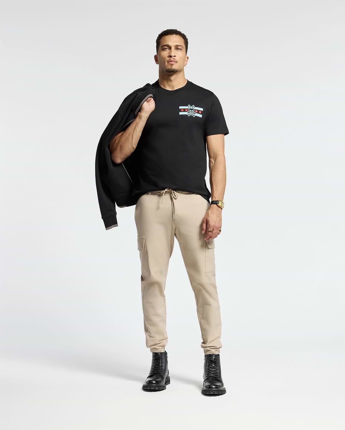 A person stands confidently against a light background, dressed in the Psycho Bunny MENS CHICAGO TEE - B6U551W1PC featuring a colorful design. They pair it with beige cargo pants and black boots, while casually holding a black jacket over one shoulder and accessorizing with a watch on their left wrist.