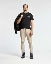 A person stands confidently against a light background, dressed in the Psycho Bunny MENS CHICAGO TEE - B6U551W1PC featuring a colorful design. They pair it with beige cargo pants and black boots, while casually holding a black jacket over one shoulder and accessorizing with a watch on their left wrist.
