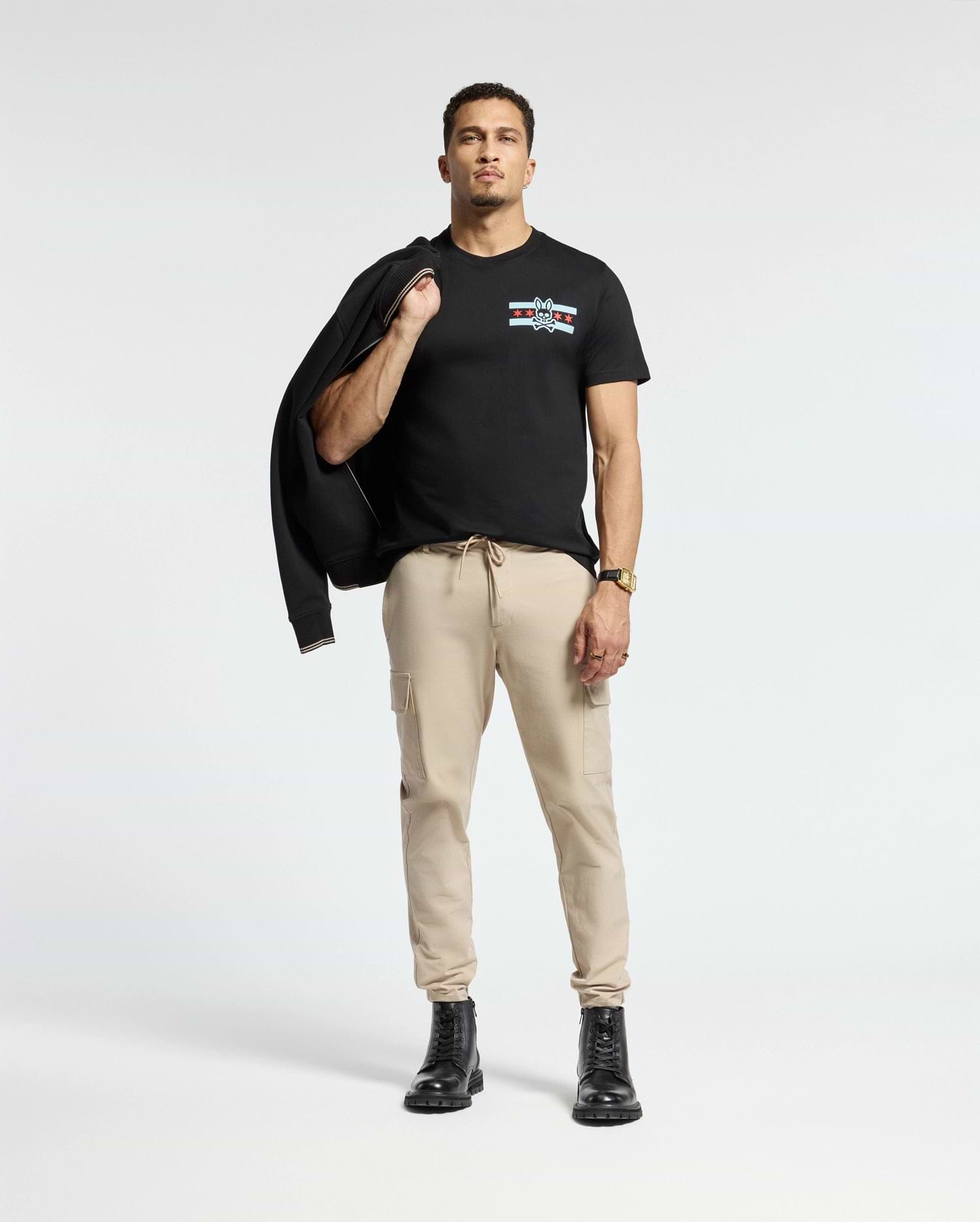 A person stands confidently against a light background, dressed in the Psycho Bunny MENS CHICAGO TEE - B6U551W1PC featuring a colorful design. They pair it with beige cargo pants and black boots, while casually holding a black jacket over one shoulder and accessorizing with a watch on their left wrist.