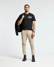 A person stands confidently against a light background, dressed in the Psycho Bunny MENS CHICAGO TEE - B6U551W1PC featuring a colorful design. They pair it with beige cargo pants and black boots, while casually holding a black jacket over one shoulder and accessorizing with a watch on their left wrist.