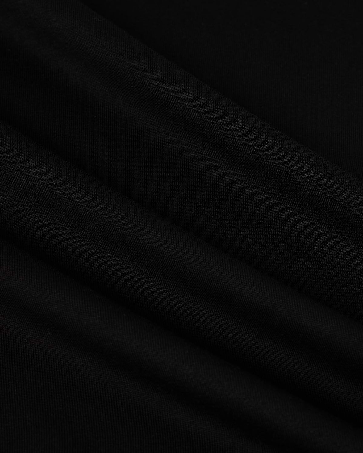A close-up image of the MENS NEW YORK TEE - B6U554W1PC by Psycho Bunny showcases its smooth, black Pima cotton fabric with soft, diagonal folds. The material has a slightly matte texture and appears to be lightweight. The folds create subtle shadows and highlights, adding depth to the otherwise uniform color. A small Bunny logo is visible in the corner.