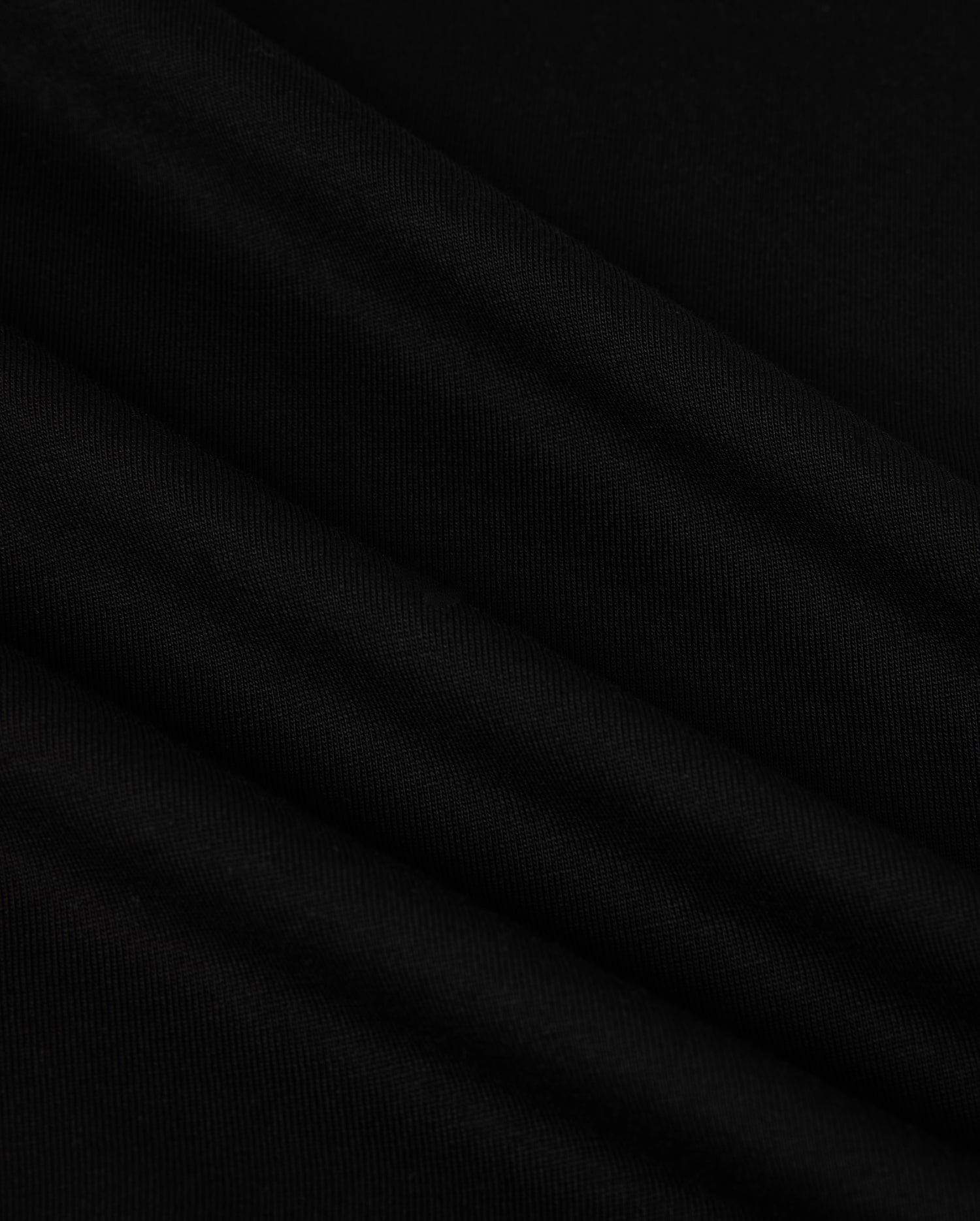 A close-up image of the MENS NEW YORK TEE - B6U554W1PC by Psycho Bunny showcases its smooth, black Pima cotton fabric with soft, diagonal folds. The material has a slightly matte texture and appears to be lightweight. The folds create subtle shadows and highlights, adding depth to the otherwise uniform color. A small Bunny logo is visible in the corner.