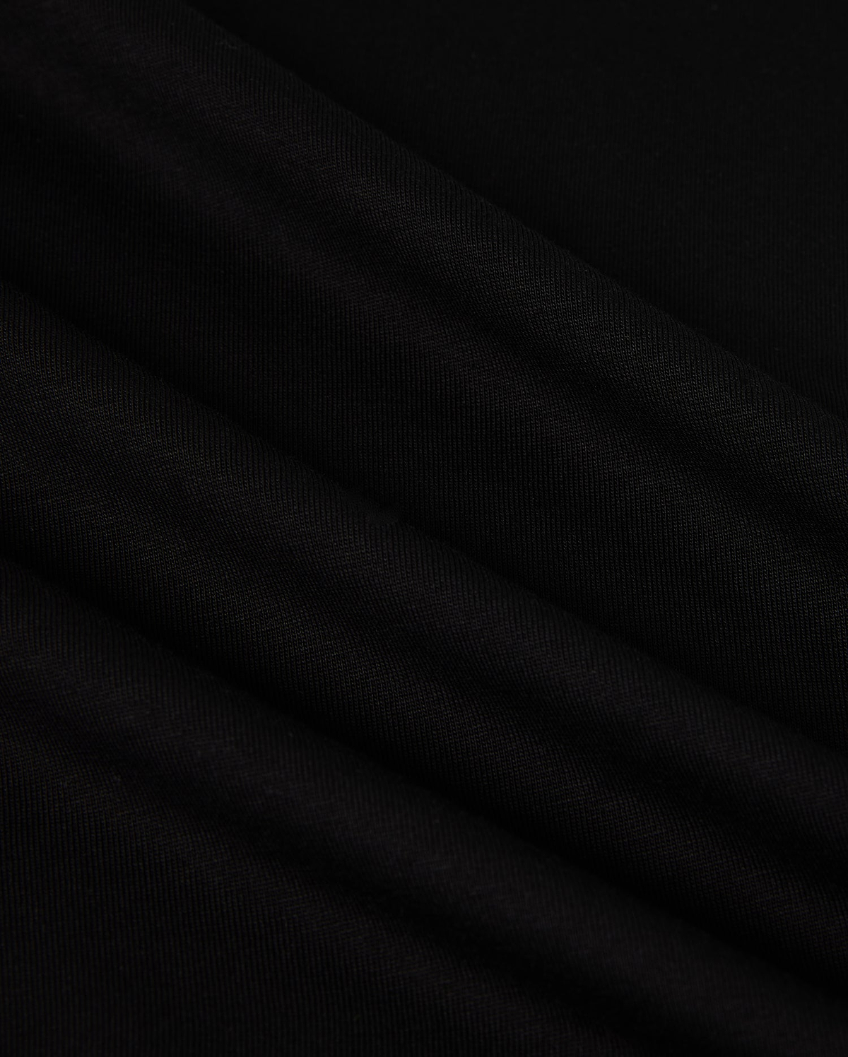 A close-up image of the MENS NEW YORK TEE - B6U554W1PC by Psycho Bunny showcases its smooth, black Pima cotton fabric with soft, diagonal folds. The material has a slightly matte texture and appears to be lightweight. The folds create subtle shadows and highlights, adding depth to the otherwise uniform color. A small Bunny logo is visible in the corner.