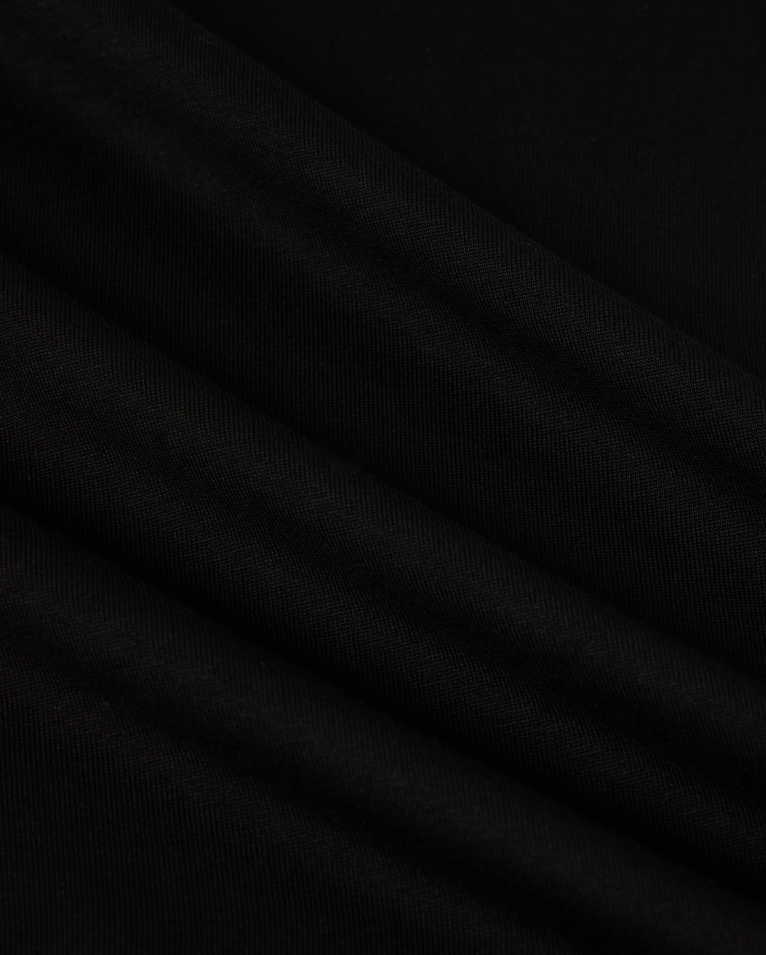 A close-up image of the MENS NEW YORK TEE - B6U554W1PC by Psycho Bunny showcases its smooth, black Pima cotton fabric with soft, diagonal folds. The material has a slightly matte texture and appears to be lightweight. The folds create subtle shadows and highlights, adding depth to the otherwise uniform color. A small Bunny logo is visible in the corner.