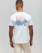 A person showcases the MENS LOS ANGELES CITY TEE (B6U861A2PC) by Psycho Bunny, featuring red line art of Los Angeles landmarks on white Pima cotton. The casual Dodgers style is complete with beige pants, complementing their short curly hair.