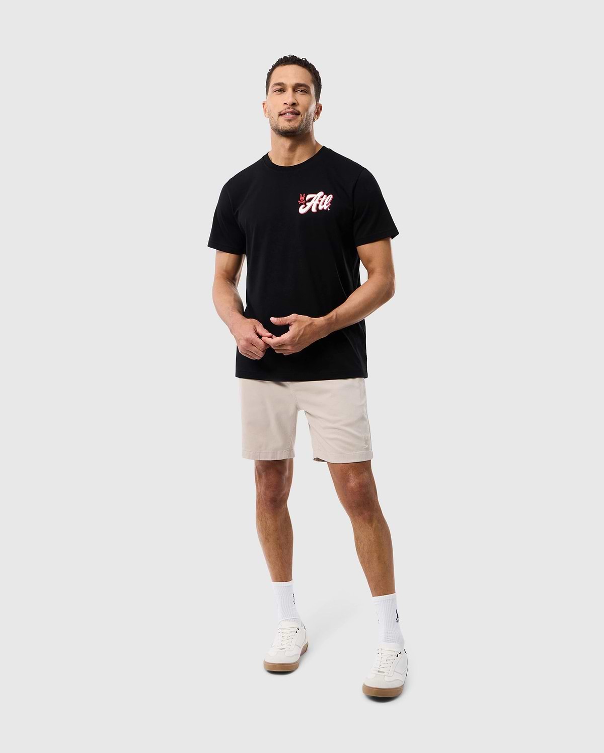 A man wearing a black Psycho Bunny ATLANTA CITY Tee with a red logo, beige shorts, and white sneakers standing against a plain white background.