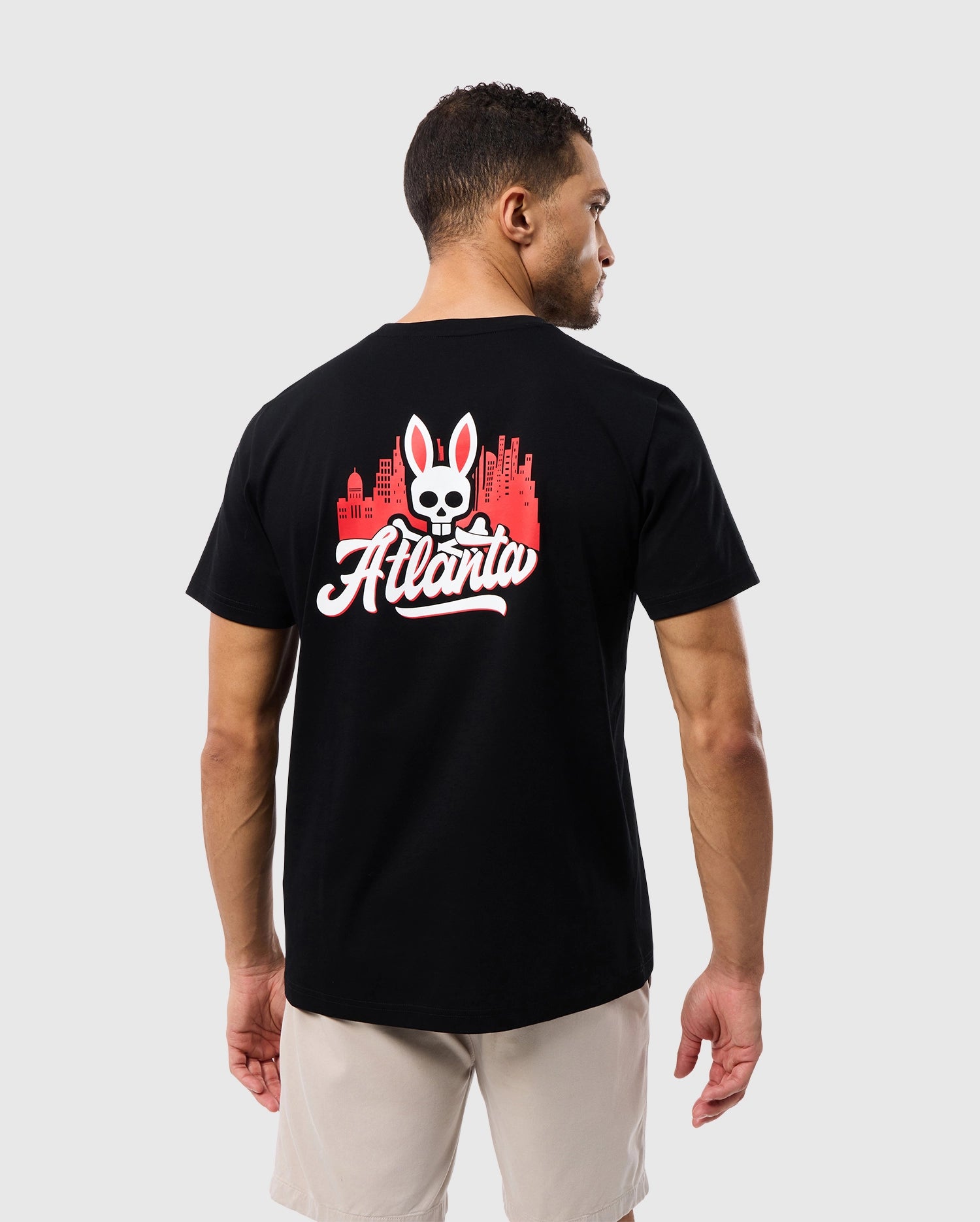 A man wearing a Psycho Bunny Black Tee crafted from Peruvian Pima cotton, featuring the MENS ATLANTA CITY TEE - B6U966X1PC graphic print with the word "Atlanta" and an emblem of a skull above crossed bones.