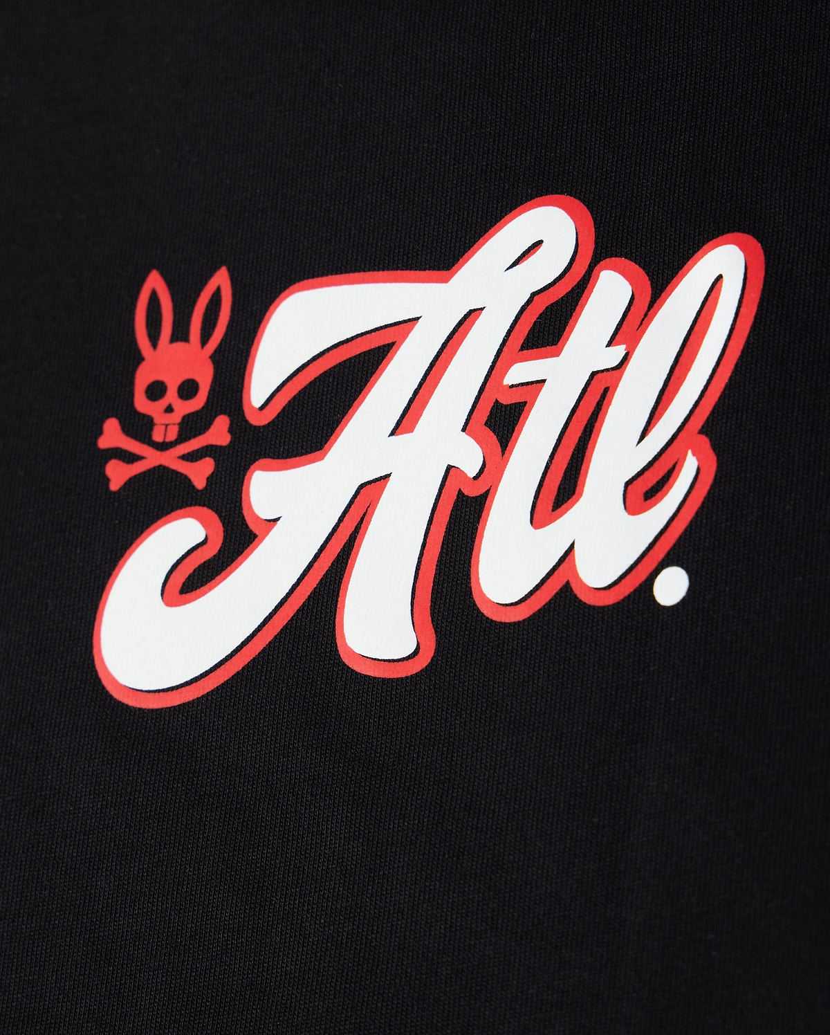 A close-up image of a white and red Psycho Bunny Atlanta City Tee logo featuring stylized script and a skull-and-crossbones symbol on a black fabric background.