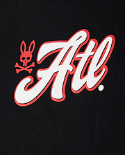 A close-up image of a white and red Psycho Bunny Atlanta City Tee logo featuring stylized script and a skull-and-crossbones symbol on a black fabric background.