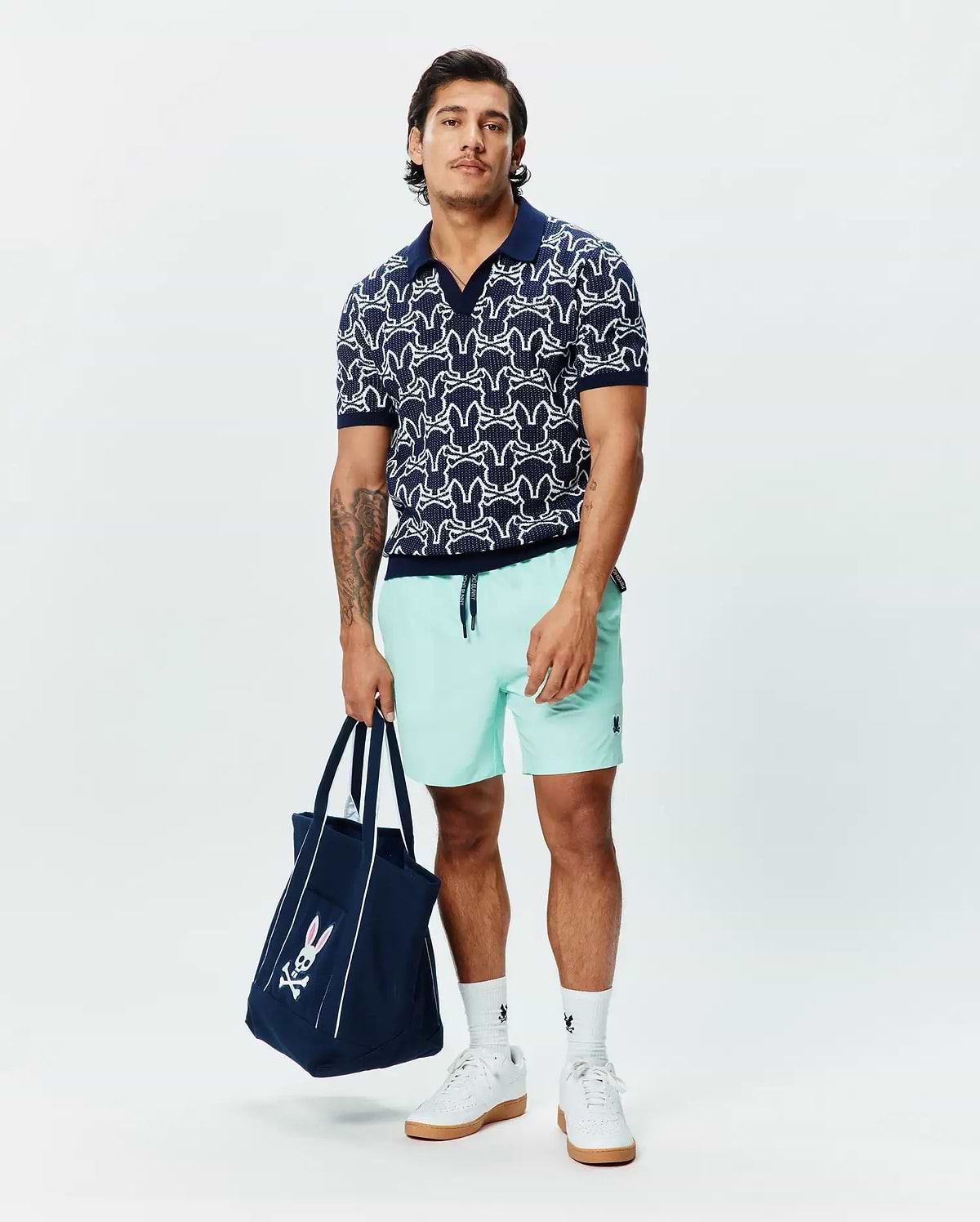 Navy knit polo with white all-over Psycho Bunny skull pattern, Mens Samos Hydrochromic Printed Swim Trunk in mint green with a small navy logo, and a navy tote bag with a large pink and white Psycho Bunny logo.