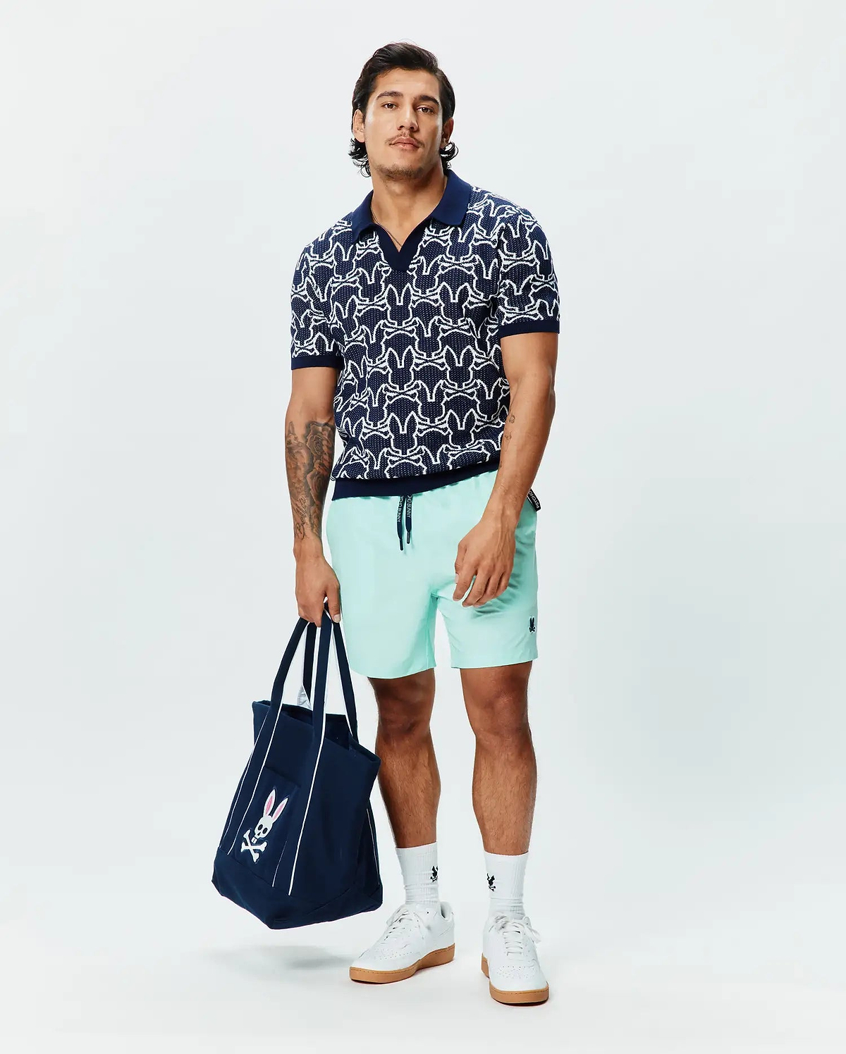 Navy knit polo with white all-over Psycho Bunny skull pattern, Mens Samos Hydrochromic Printed Swim Trunk in mint green with a small navy logo, and a navy tote bag with a large pink and white Psycho Bunny logo.