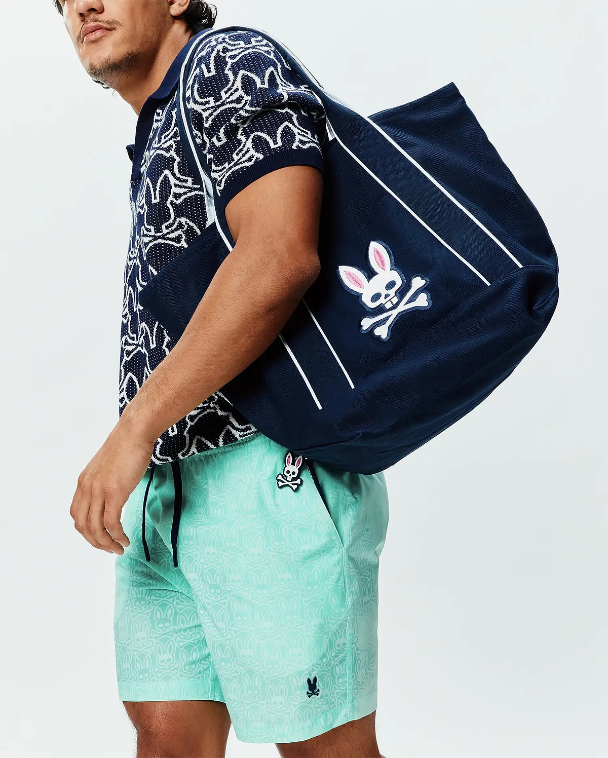 Navy textured knit polo with white abstract bunny pattern, Mens Samos Hydrochromic Printed Swim Trunk 6 in mint green with tonal bunny skull print and small embroidered logo, plus a navy tote with large pink-eared bunny skull logo.