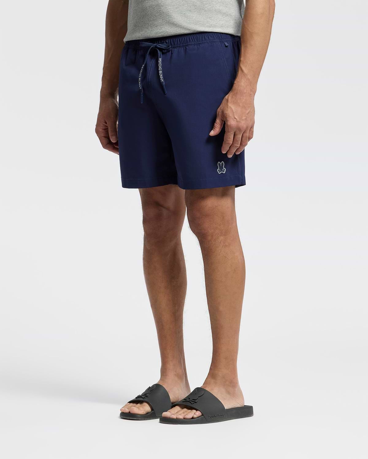 A person wearing a grey t-shirt, Psycho Bunny MENS SAMOS HYDROCHROMIC PRINTED SWIM TRUNK - B6W453F200 with a white bunny logo, and black slide sandals against a plain light background.