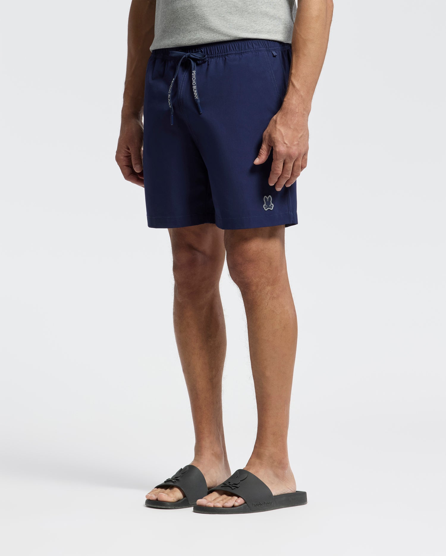 A person wearing a grey t-shirt, Psycho Bunny MENS SAMOS HYDROCHROMIC PRINTED SWIM TRUNK - B6W453F200 with a white bunny logo, and black slide sandals against a plain light background.