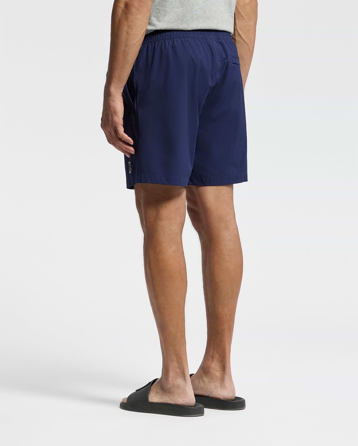 A person stands facing away from the camera against a white background, wearing a light gray T-shirt, black slide sandals, and Psycho Bunny MENS SAMOS HYDROCHROMIC PRINTED SWIM TRUNK (B6W453F200).