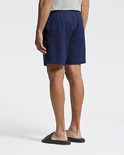A person stands facing away from the camera against a white background, wearing a light gray T-shirt, black slide sandals, and Psycho Bunny MENS SAMOS HYDROCHROMIC PRINTED SWIM TRUNK (B6W453F200).