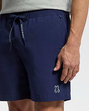 A person wears Psycho Bunny’s MENS SAMOS HYDROCHROMIC PRINTED SWIM TRUNK (B6W453F200) in navy blue, featuring a small bunny logo on the left leg and "Psycho Bunny" text on the drawstring; only torso and upper legs are visible.