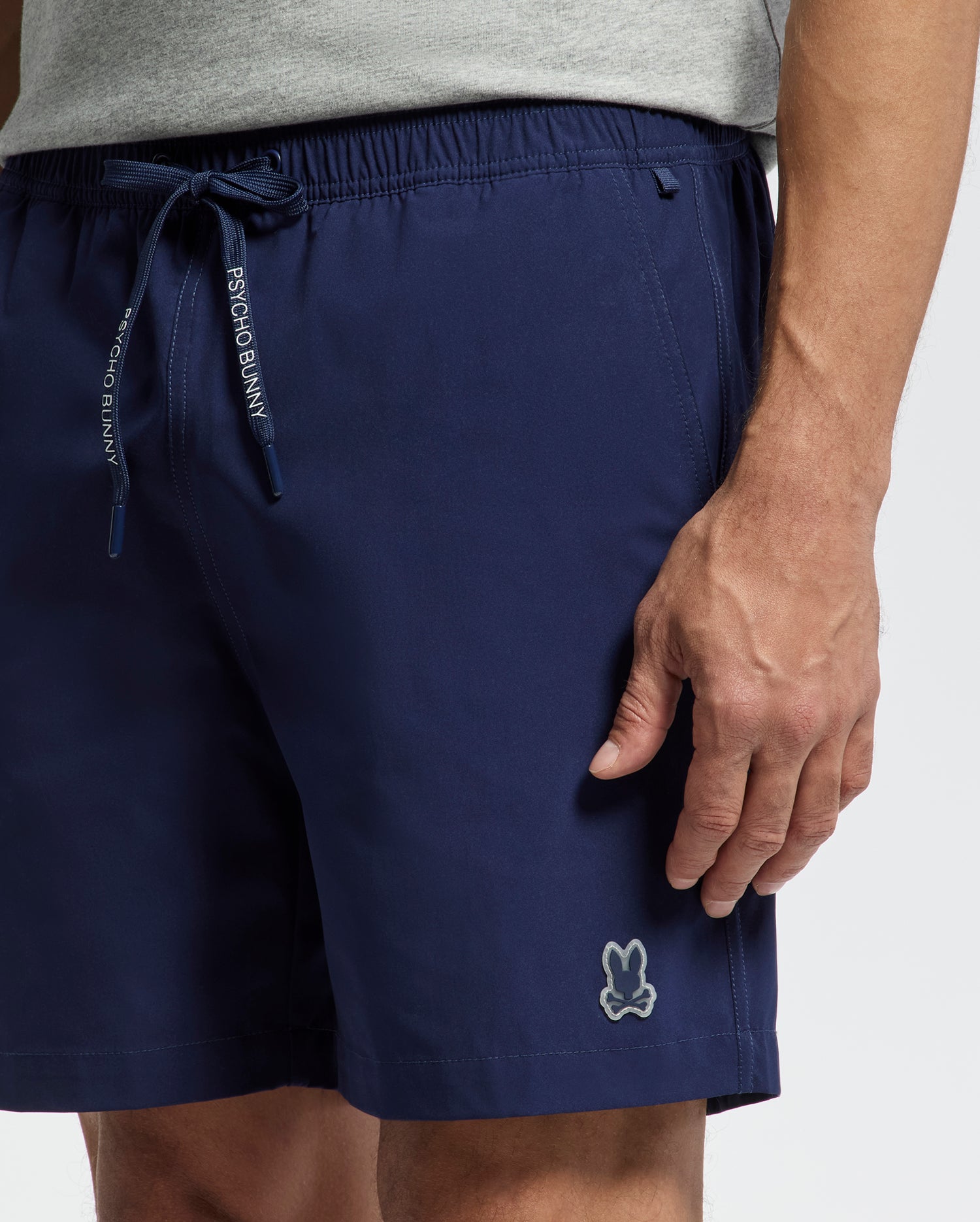 A person wears Psycho Bunny’s MENS SAMOS HYDROCHROMIC PRINTED SWIM TRUNK (B6W453F200) in navy blue, featuring a small bunny logo on the left leg and "Psycho Bunny" text on the drawstring; only torso and upper legs are visible.
