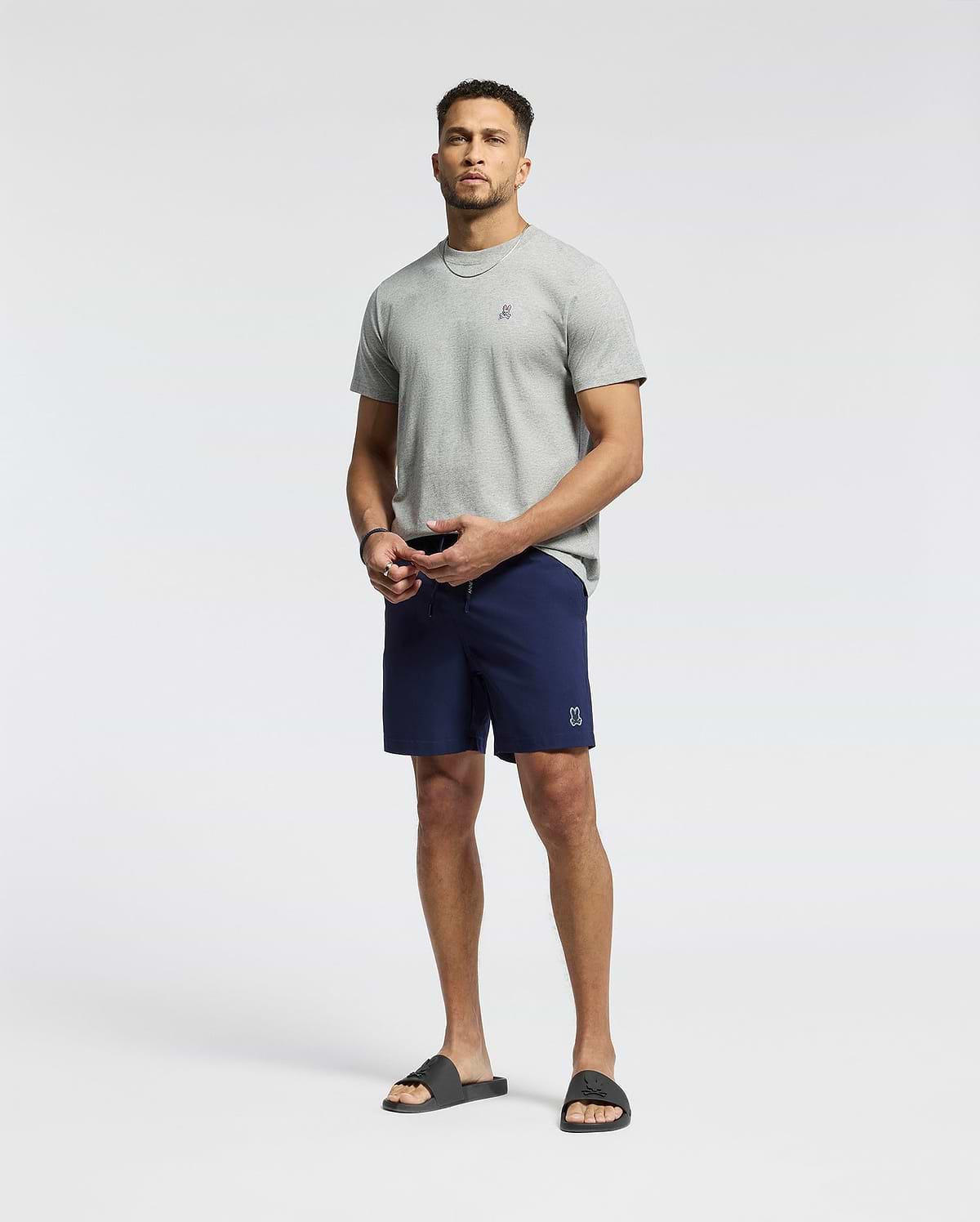 A man stands against a plain background wearing a light gray T-shirt and Psycho Bunny MENS SAMOS HYDROCHROMIC PRINTED SWIM TRUNK. He completes the look with black slide sandals, has a short beard, and gazes at the camera neutrally.