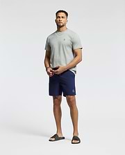 A man stands against a plain background wearing a light gray T-shirt and Psycho Bunny MENS SAMOS HYDROCHROMIC PRINTED SWIM TRUNK. He completes the look with black slide sandals, has a short beard, and gazes at the camera neutrally.