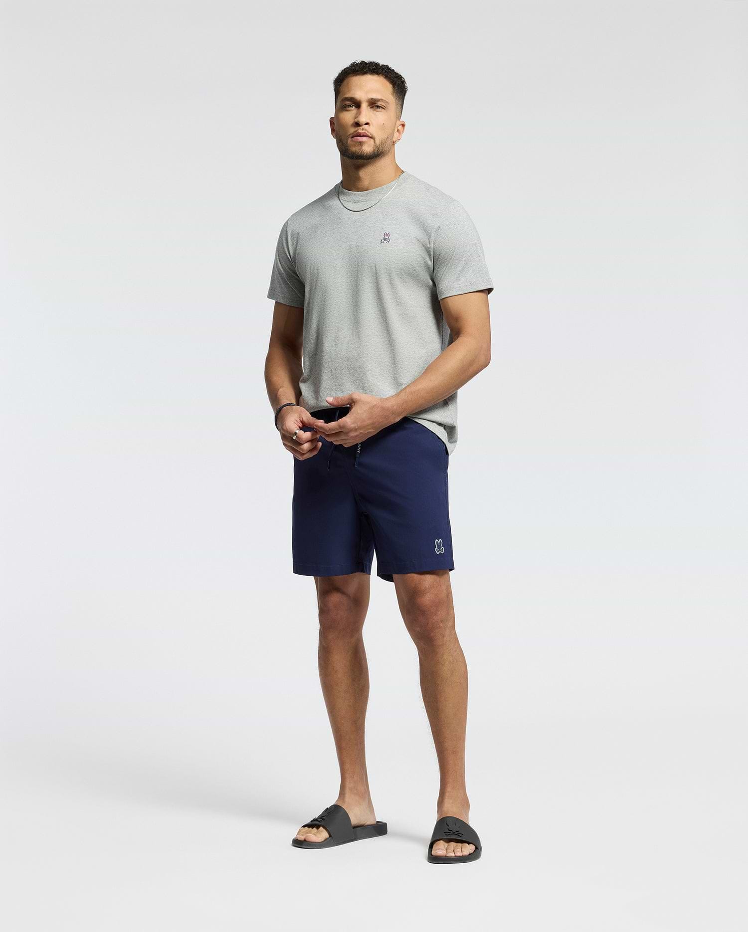 A man stands against a plain background wearing a light gray T-shirt and Psycho Bunny MENS SAMOS HYDROCHROMIC PRINTED SWIM TRUNK. He completes the look with black slide sandals, has a short beard, and gazes at the camera neutrally.