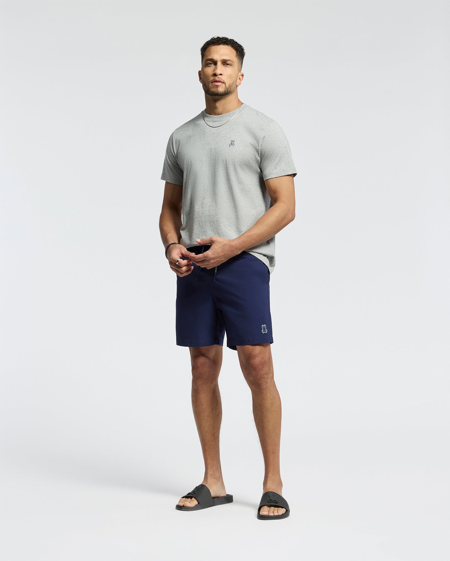 A man stands against a plain background wearing a light gray T-shirt and Psycho Bunny MENS SAMOS HYDROCHROMIC PRINTED SWIM TRUNK. He completes the look with black slide sandals, has a short beard, and gazes at the camera neutrally.