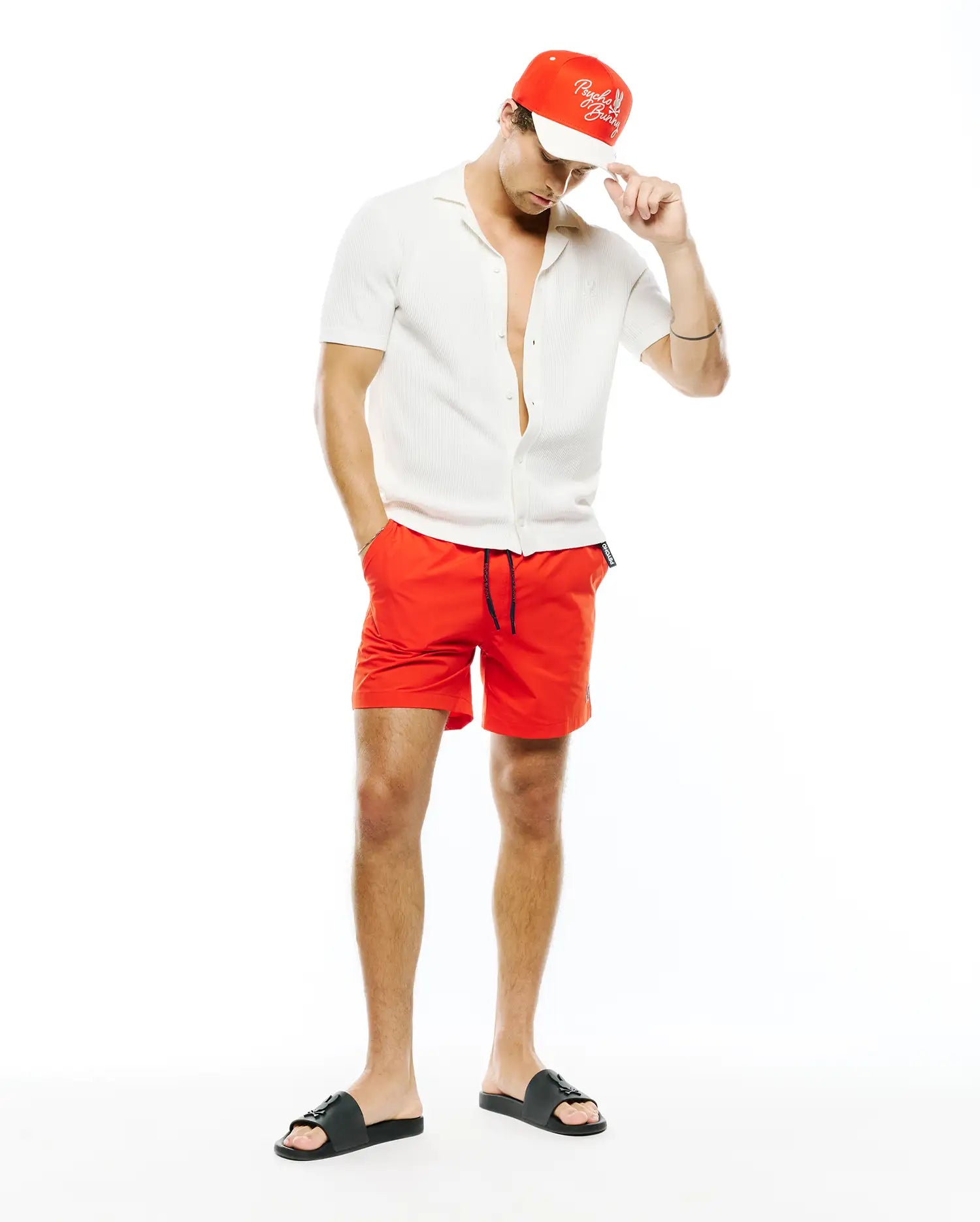 Mens Samos Hydrochromic Printed Swim Trunk in vibrant red with black drawstring, a white ribbed knit short-sleeve collared shirt, a red Psycho Bunny script logo cap, and black logo slides.