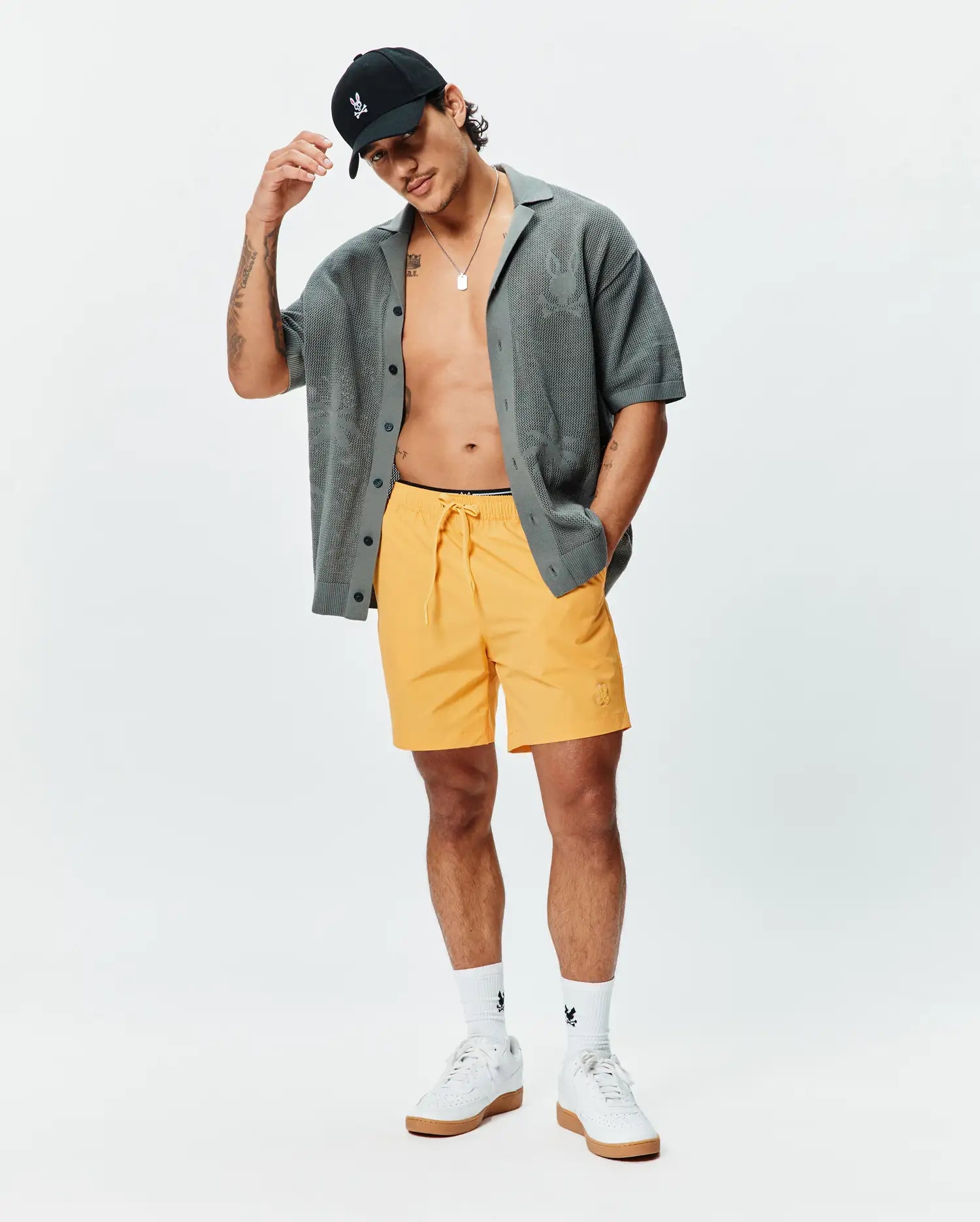 Mens Samos Hydrochromic Printed Swim Trunk in bright yellow with an embroidered Psycho Bunny skull logo, a relaxed charcoal knit short-sleeve shirt with subtle skull pattern, and a black cap with an embroidered skull. (5)