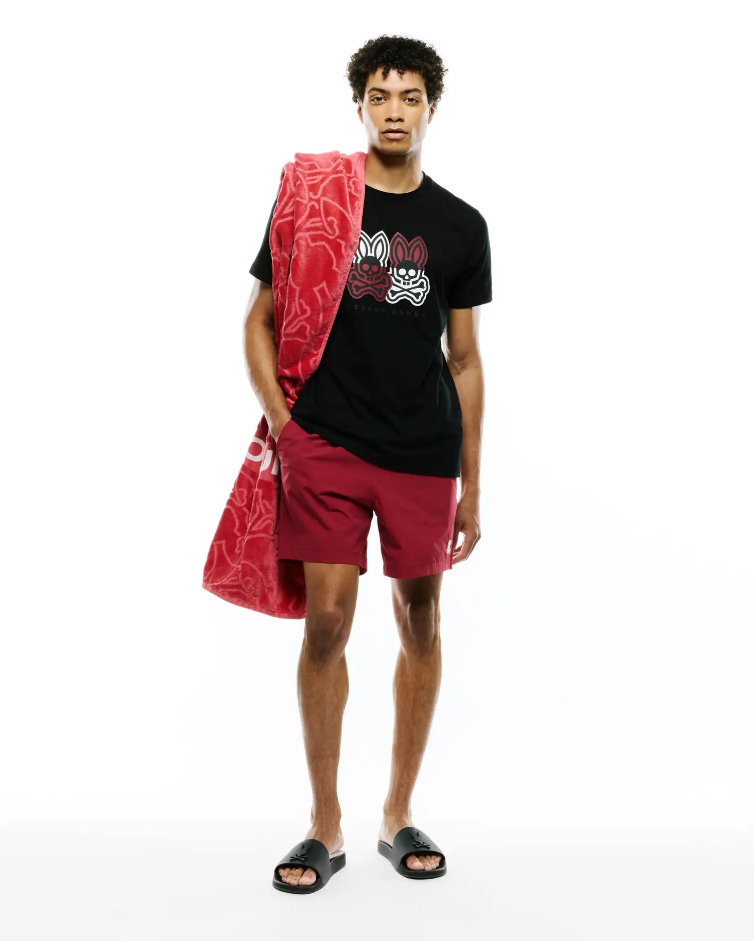 Black jersey t-shirt with large red, white, and black Psycho Bunny skull graphic on chest, paired with Mens Samos Hydrochromic Printed Swim Trunk in maroon with a small white logo, and a red patterned terry towel.