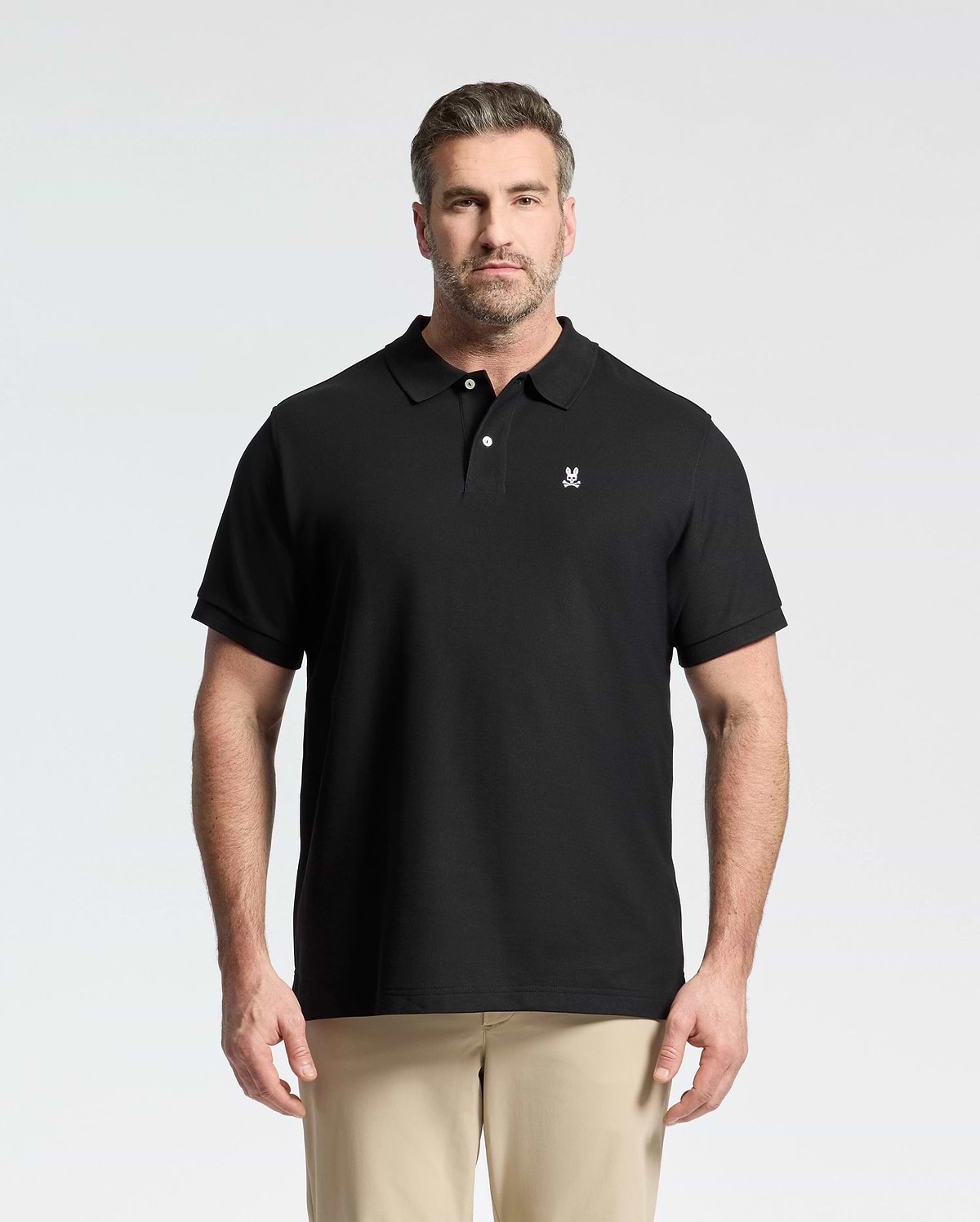 A man with short hair and a beard wears the Psycho Bunny Men's Big and Tall Classic Pique Polo Shirt (B9K001ARPC) in black with a small white logo, paired with beige pants, standing against a plain light background.