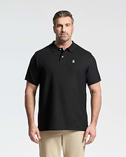 A man with short hair and a beard wears the Psycho Bunny Men's Big and Tall Classic Pique Polo Shirt (B9K001ARPC) in black with a small white logo, paired with beige pants, standing against a plain light background.