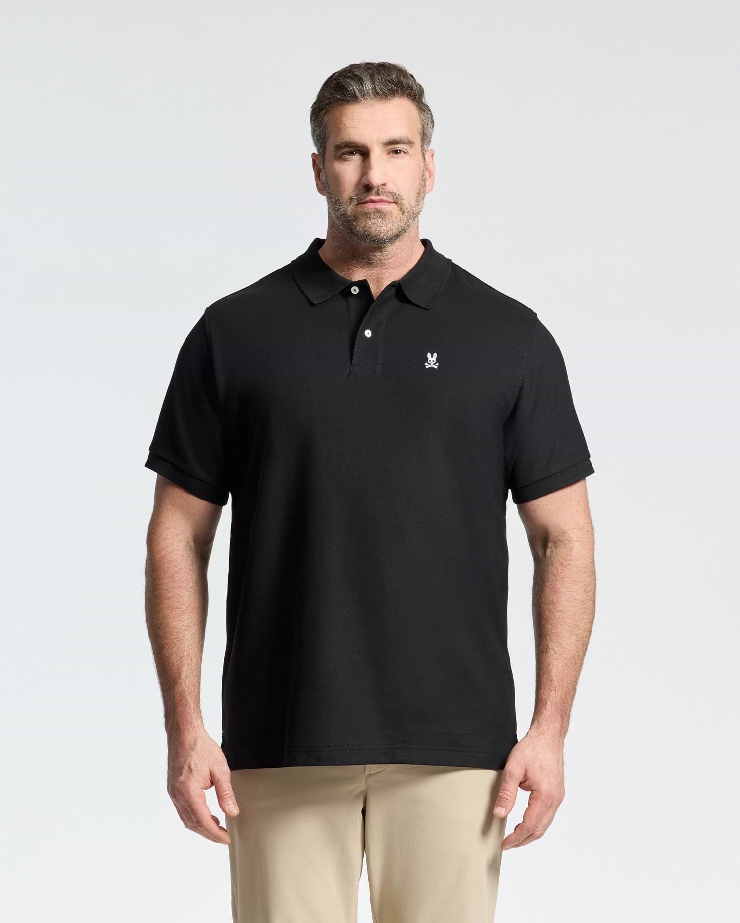 A man with short hair and a beard wears the Psycho Bunny Men's Big and Tall Classic Pique Polo Shirt (B9K001ARPC) in black with a small white logo, paired with beige pants, standing against a plain light background.
