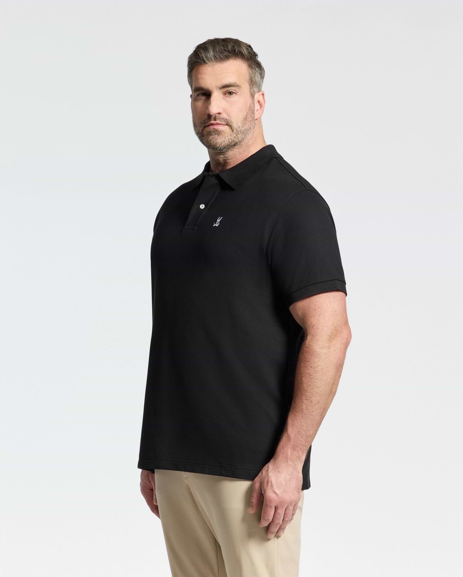 A man with short brown hair and a beard wears a Psycho Bunny MENS BIG AND TALL CLASSIC PIQUE POLO SHIRT - B9K001ARPC in black and beige pants, standing against a plain white background and looking slightly to the side.