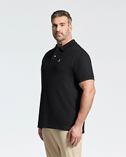 A man with short brown hair and a beard wears a Psycho Bunny MENS BIG AND TALL CLASSIC PIQUE POLO SHIRT - B9K001ARPC in black and beige pants, standing against a plain white background and looking slightly to the side.