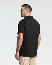 A man with short hair and a beard stands facing away, wearing the Psycho Bunny MENS BIG AND TALL CLASSIC PIQUE POLO SHIRT - B9K001ARPC in black Pima cotton and beige pants against a plain white background.