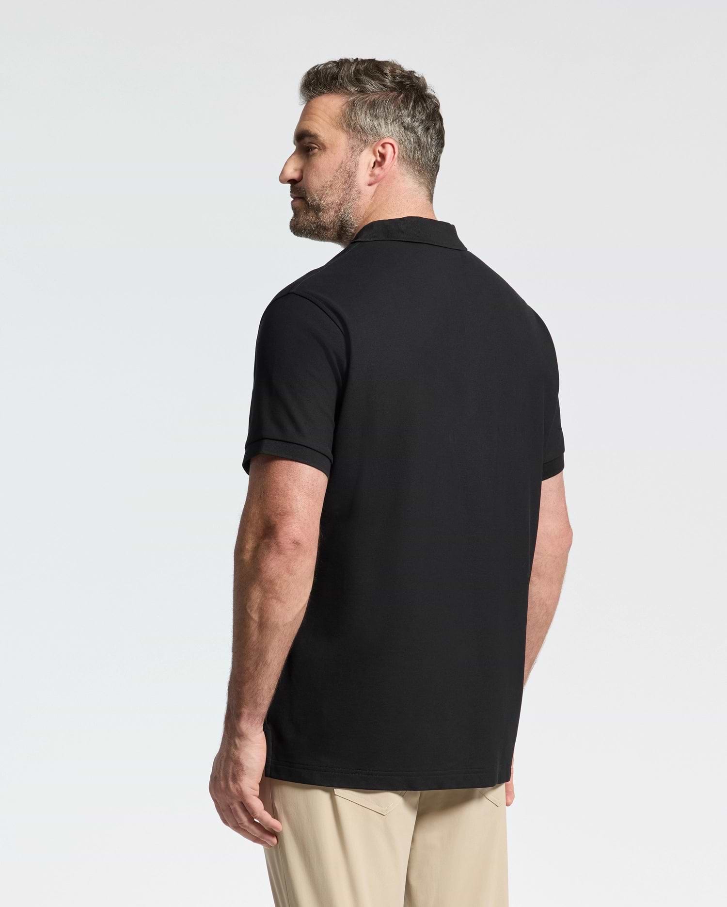 A man with short hair and a beard stands facing away, wearing the Psycho Bunny MENS BIG AND TALL CLASSIC PIQUE POLO SHIRT - B9K001ARPC in black Pima cotton and beige pants against a plain white background.