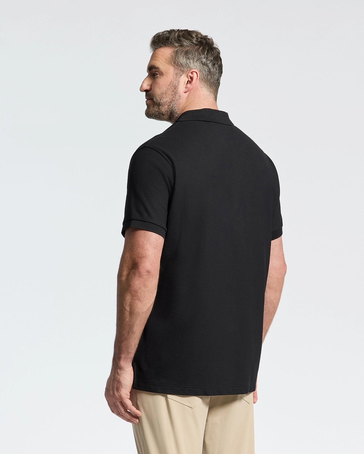 A man with short hair and a beard stands facing away, wearing the Psycho Bunny MENS BIG AND TALL CLASSIC PIQUE POLO SHIRT - B9K001ARPC in black Pima cotton and beige pants against a plain white background.