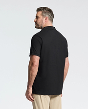 A man with short hair and a beard stands facing away, wearing the Psycho Bunny MENS BIG AND TALL CLASSIC PIQUE POLO SHIRT - B9K001ARPC in black Pima cotton and beige pants against a plain white background.