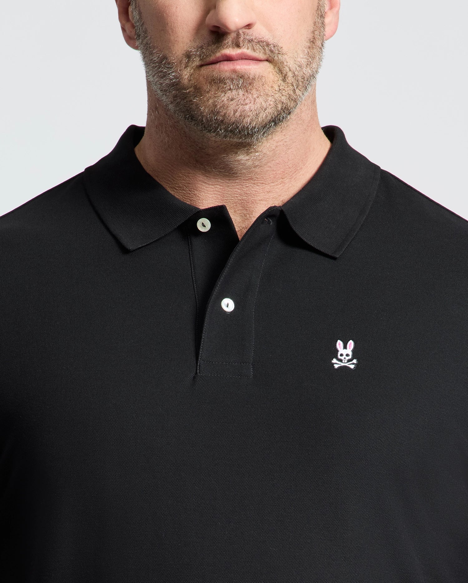 A man, visible from the lower face up, wears a Psycho Bunny MENS BIG AND TALL CLASSIC PIQUE POLO SHIRT—black Pima cotton with a small white and pink skull and bunny ears logo on the left chest.