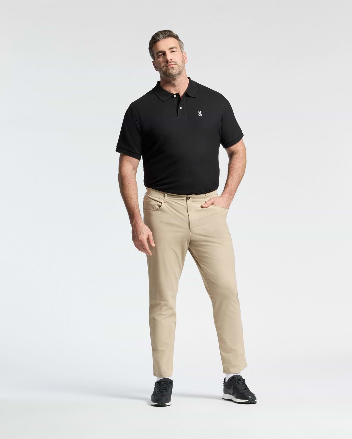 A man poses against a white background wearing a Psycho Bunny MENS BIG AND TALL CLASSIC PIQUE POLO SHIRT - B9K001ARPC, paired with beige pants and black sneakers. One hand is in his pocket, the other by his side.