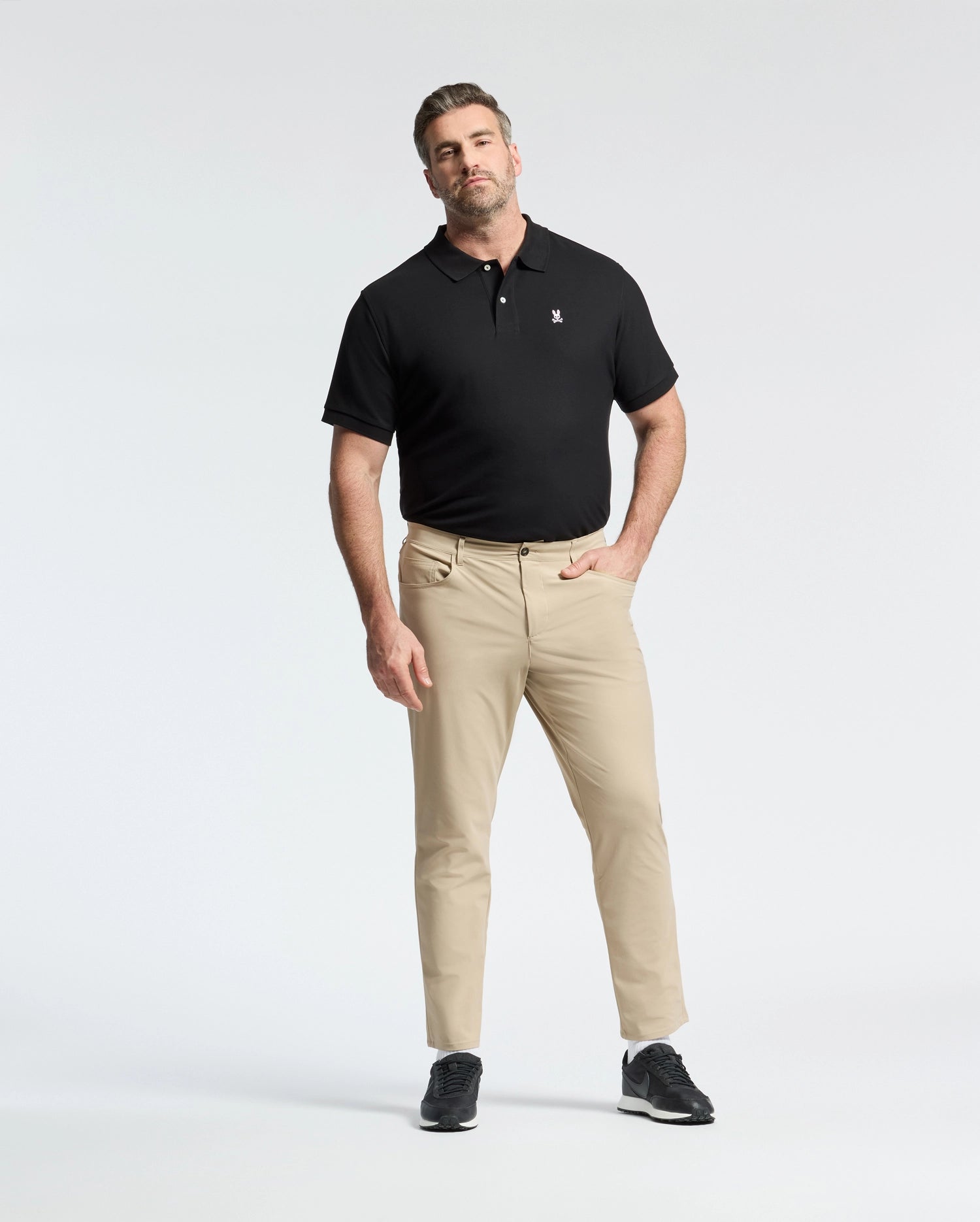 A man poses against a white background wearing a Psycho Bunny MENS BIG AND TALL CLASSIC PIQUE POLO SHIRT - B9K001ARPC, paired with beige pants and black sneakers. One hand is in his pocket, the other by his side.
