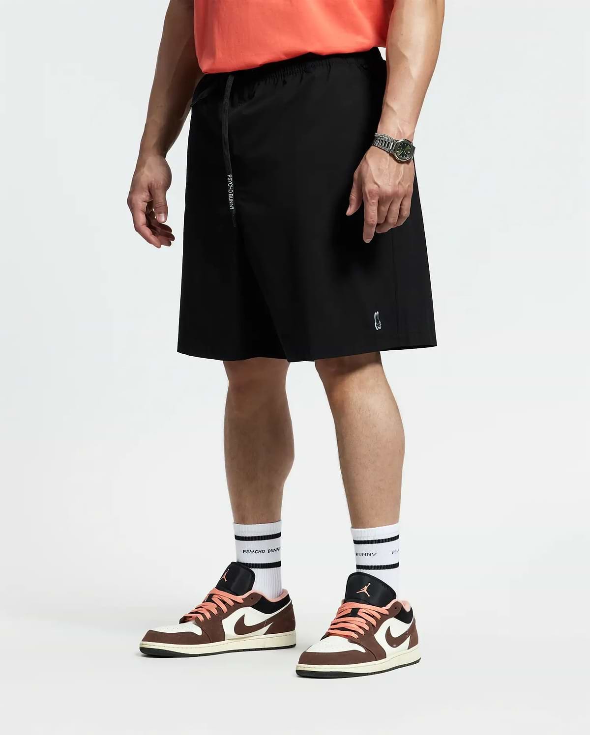 A person in coral quick-dry t-shirt, white socks, brown and white sneakers, and black Psycho Bunny MENS BIG AND TALL SAMOS HYDROCHROMIC SWIM TRUNK (B9W453F200) stands facing sideways with a watch on their relaxed left wrist.