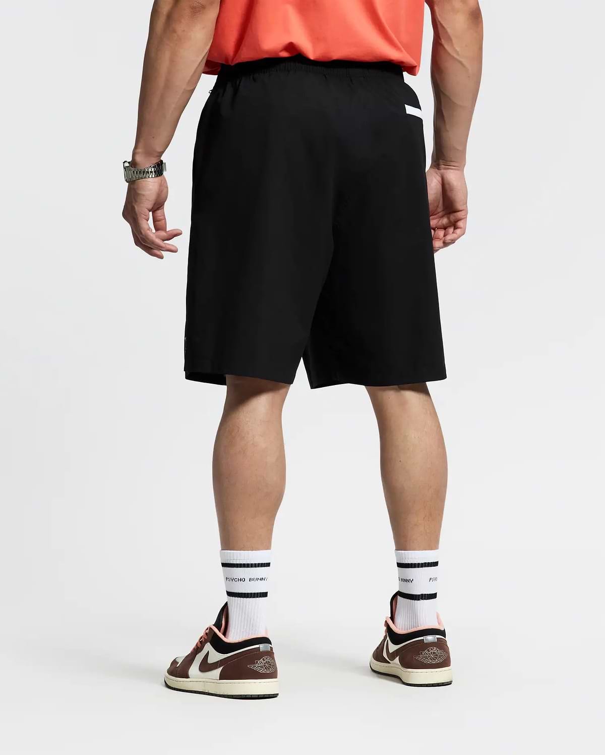A person faces away, wearing a red shirt; Psycho Bunny MENS BIG AND TALL SAMOS HYDROCHROMIC SWIM TRUNK (black, quick-dry); white socks with black stripes; brown and white sneakers; a watch on the left wrist. The background is plain and light.