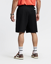 A person faces away, wearing a red shirt; Psycho Bunny MENS BIG AND TALL SAMOS HYDROCHROMIC SWIM TRUNK (black, quick-dry); white socks with black stripes; brown and white sneakers; a watch on the left wrist. The background is plain and light.