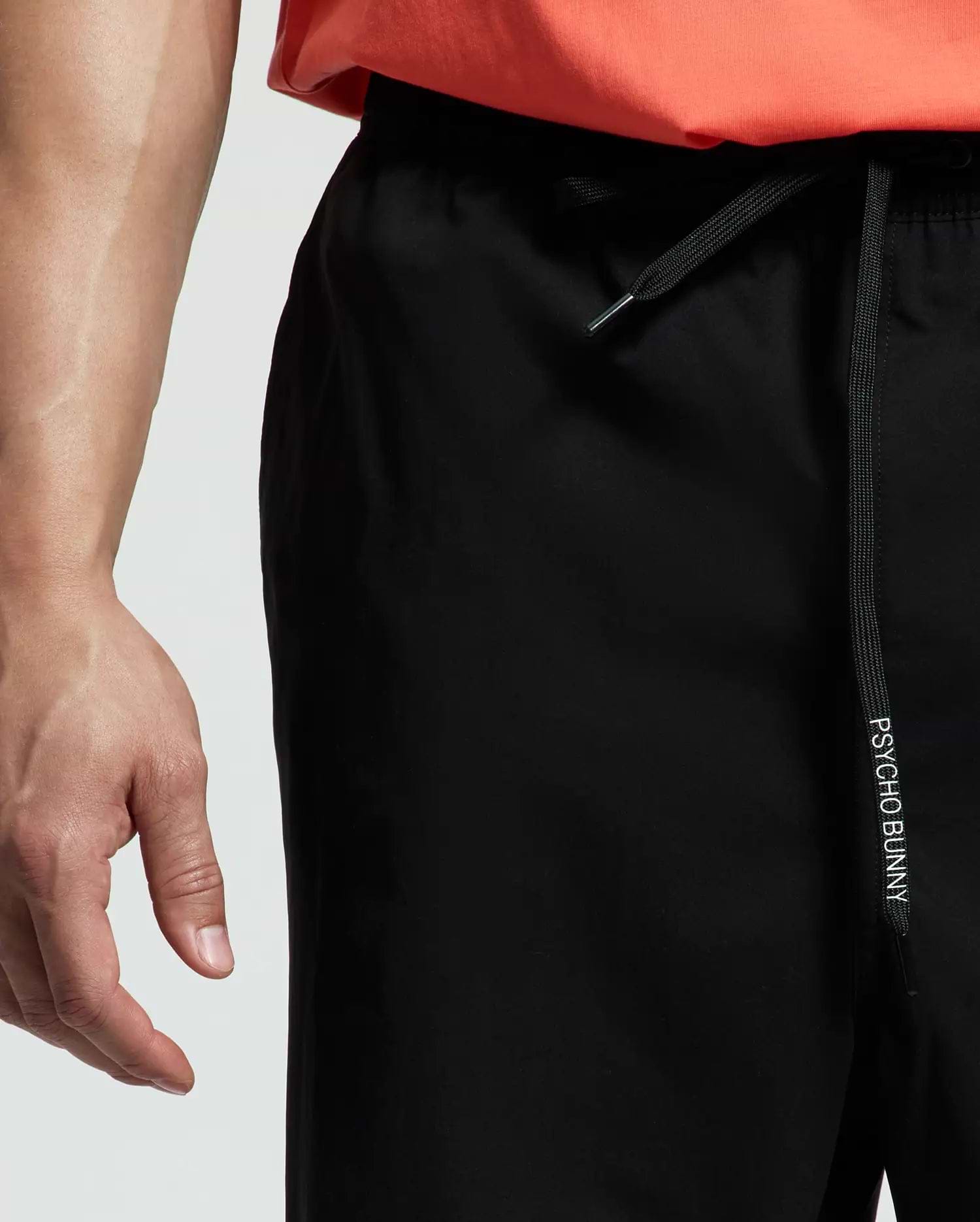 A person wears Psycho Bunny MENS BIG AND TALL SAMOS HYDROCHROMIC SWIM TRUNK with "PSYCHO BUNNY" on the drawstring and a coral shirt. The image highlights the waist area and part of the hand.