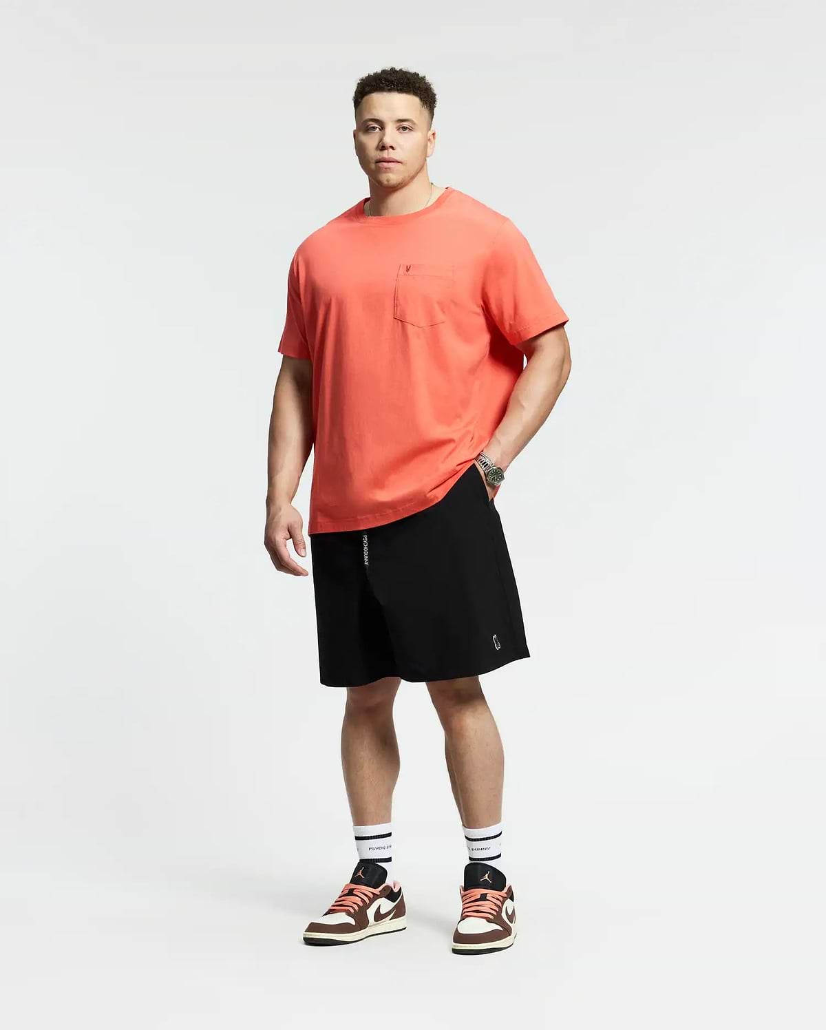 A man faces forward against a white background, wearing a coral t-shirt with a chest pocket, Psycho Bunny MENS BIG AND TALL SAMOS HYDROCHROMIC SWIM TRUNK (B9W453F200), white socks with black stripes, and red-and-white sneakers.