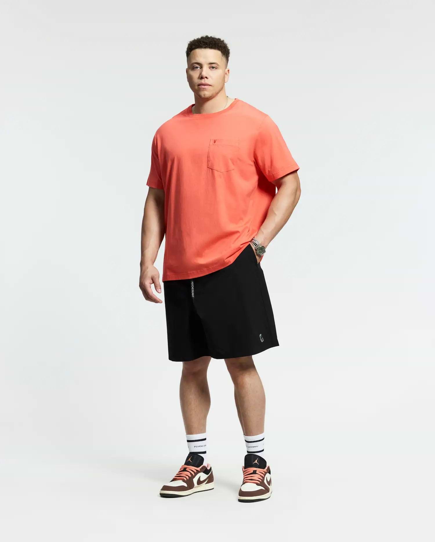 A man faces forward against a white background, wearing a coral t-shirt with a chest pocket, Psycho Bunny MENS BIG AND TALL SAMOS HYDROCHROMIC SWIM TRUNK (B9W453F200), white socks with black stripes, and red-and-white sneakers.
