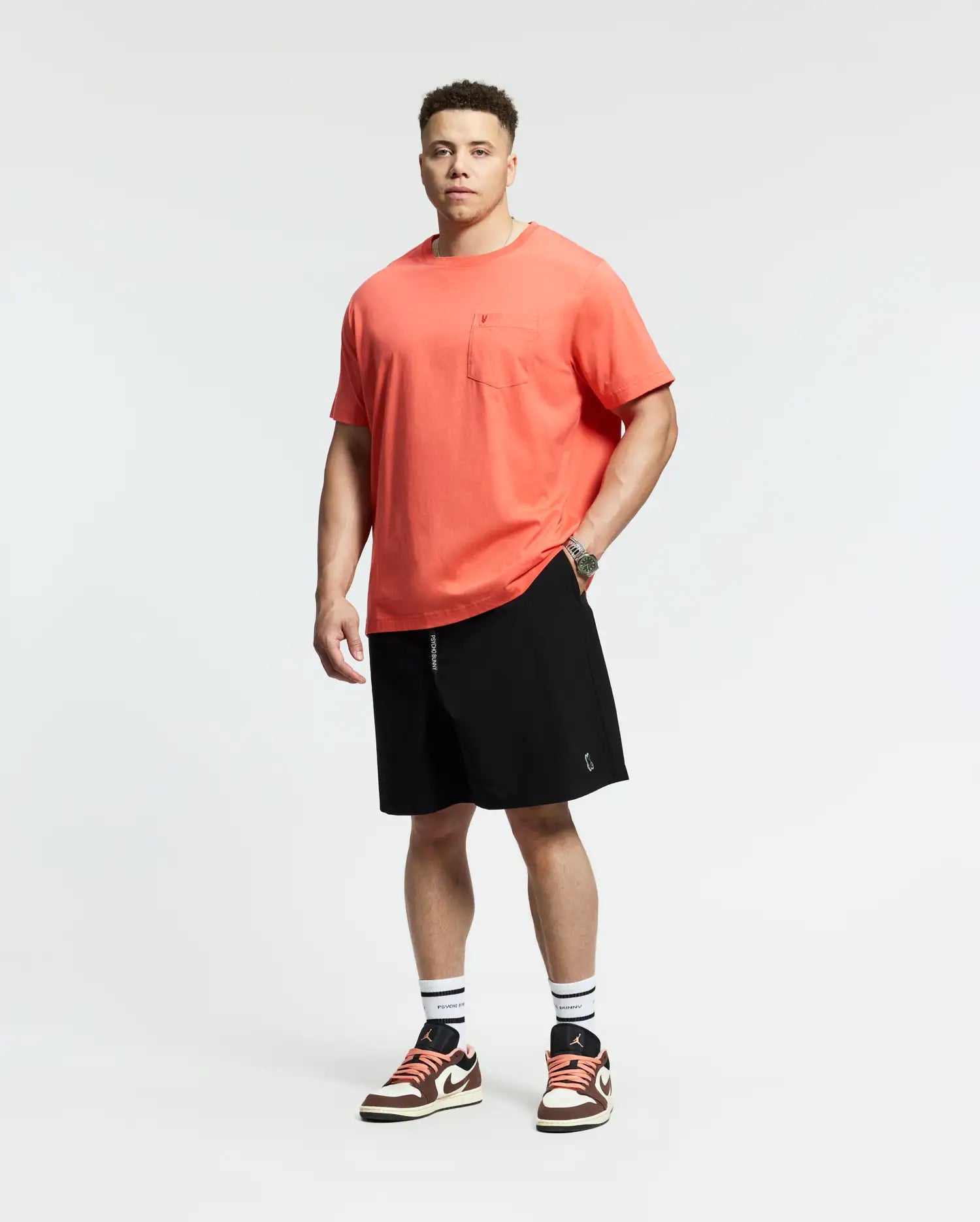 A man faces forward against a white background, wearing a coral t-shirt with a chest pocket, Psycho Bunny MENS BIG AND TALL SAMOS HYDROCHROMIC SWIM TRUNK (B9W453F200), white socks with black stripes, and red-and-white sneakers.